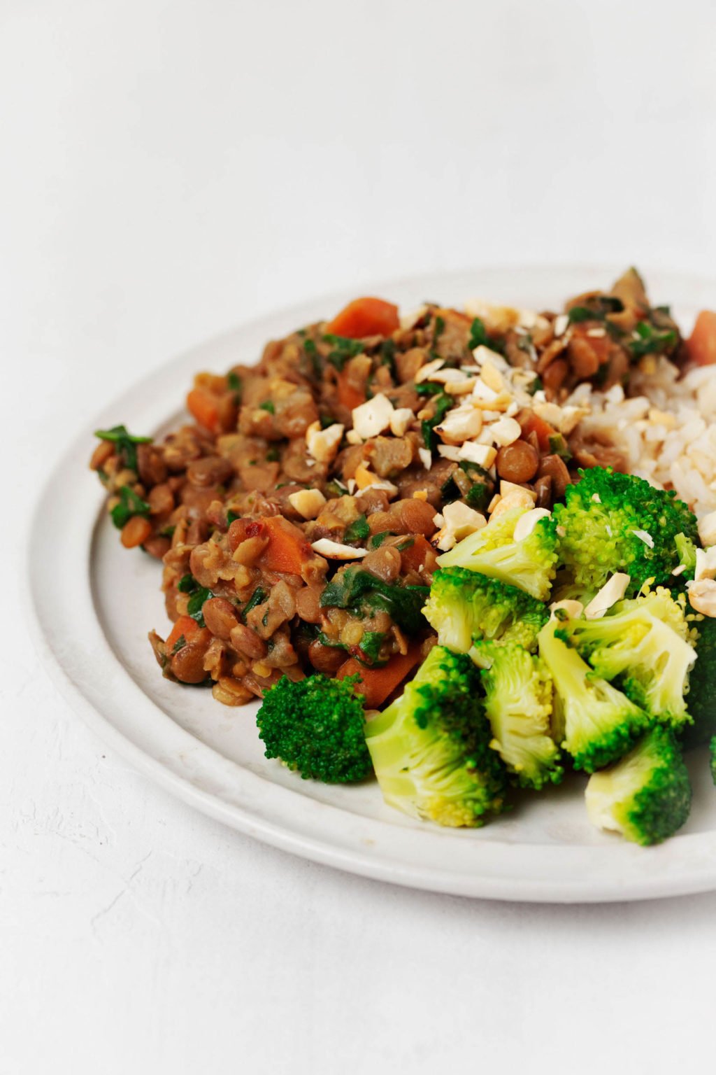 A white plate has been piled high with bright green broccoli, lentils, rice, and chopped nuts. It's resting on a white surface.