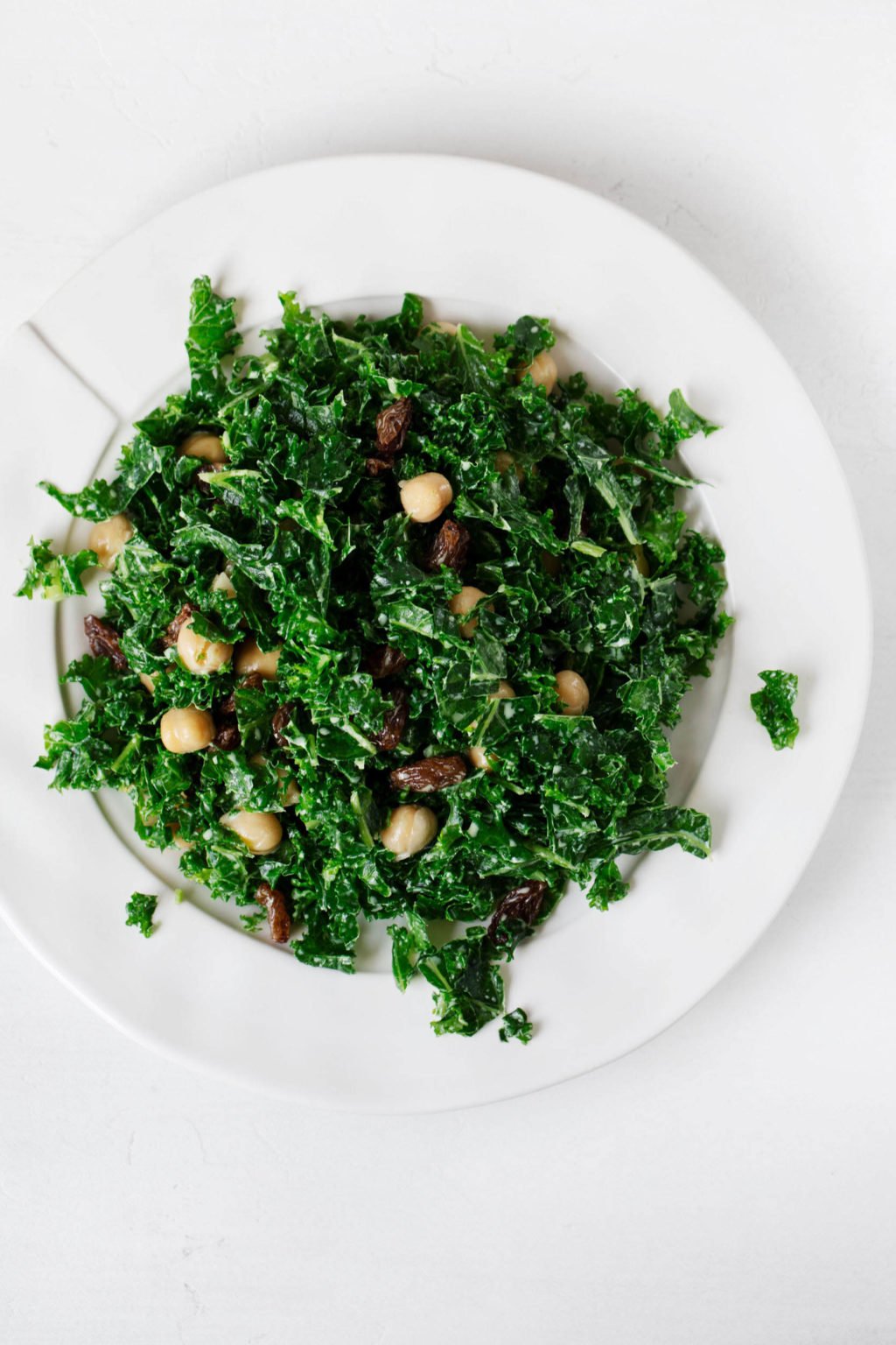 A white, rimmed salad plate has been piled with kale, chickpeas, raisins, and a creamy, tahini-based dressing.