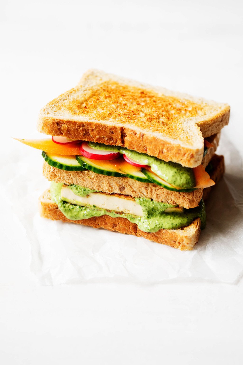 A colorful, three layered vegan green goddess sandwich is placed on a small piece of parchment paper, waiting to be cut in half.