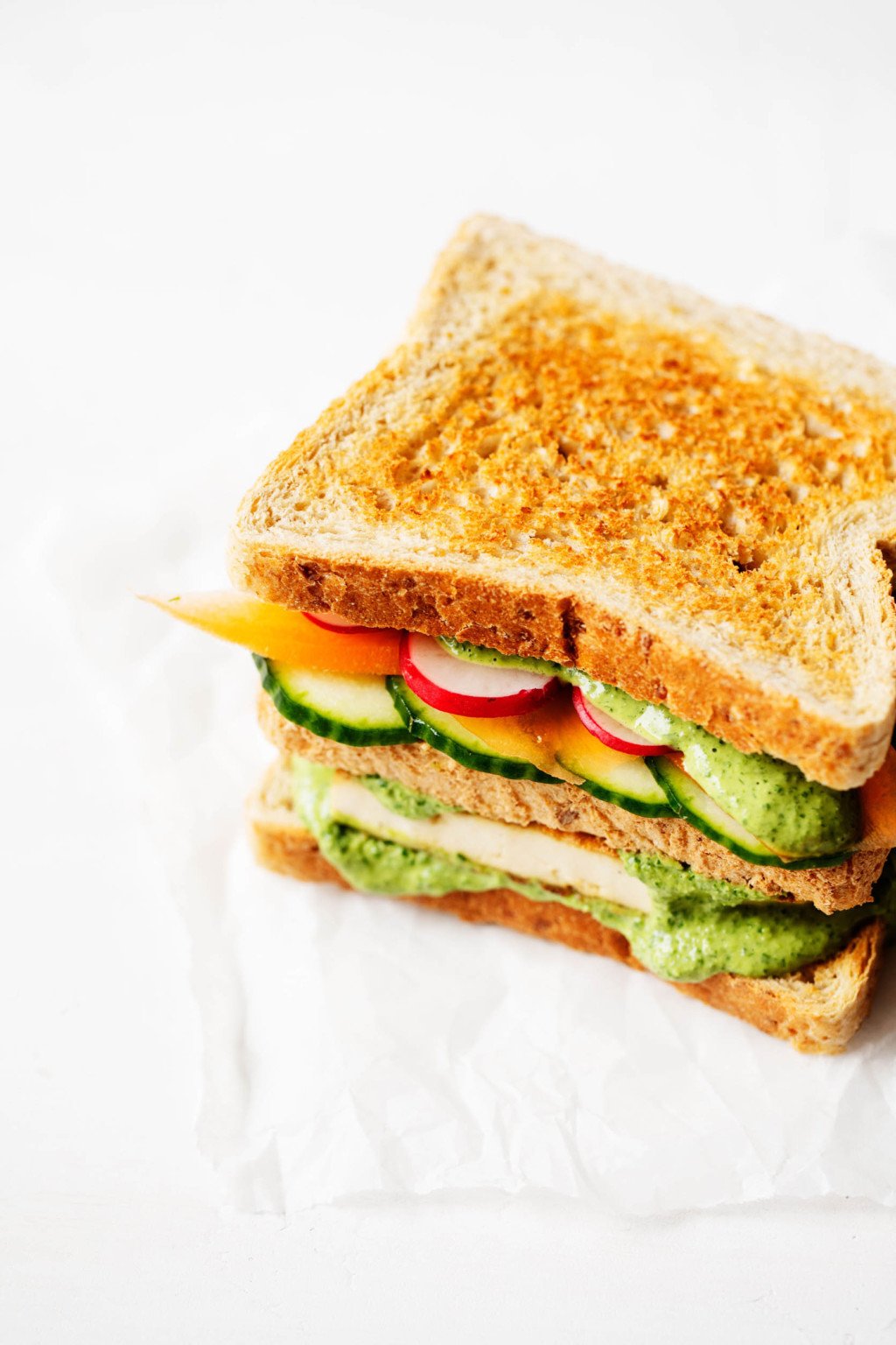 A three layered, plant based sandwich with vegetables and tofu is placed on a small piece of parchment.