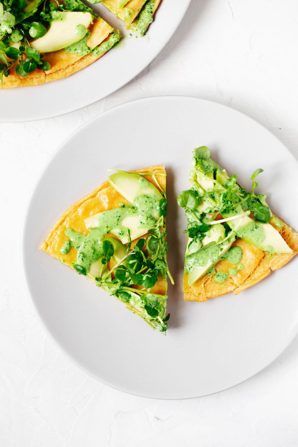 Two small slices of a chickpea flatbread, topped with avocado and greens, are on a white serving plate. 