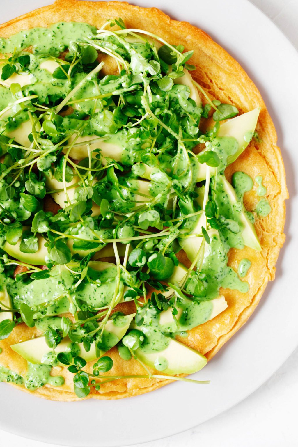 A close up, overhead image of a vegan socca flatbread, which is smothered in a bright herb sauce and vegetables.