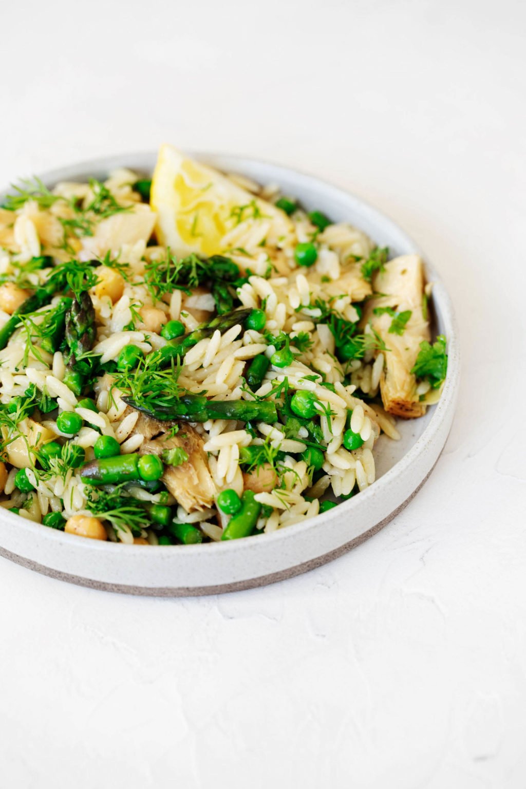 A rimmed, round white plate has been filled with an orzo pasta salad that's packed with vegetables and herbs. It rests on a white surface.