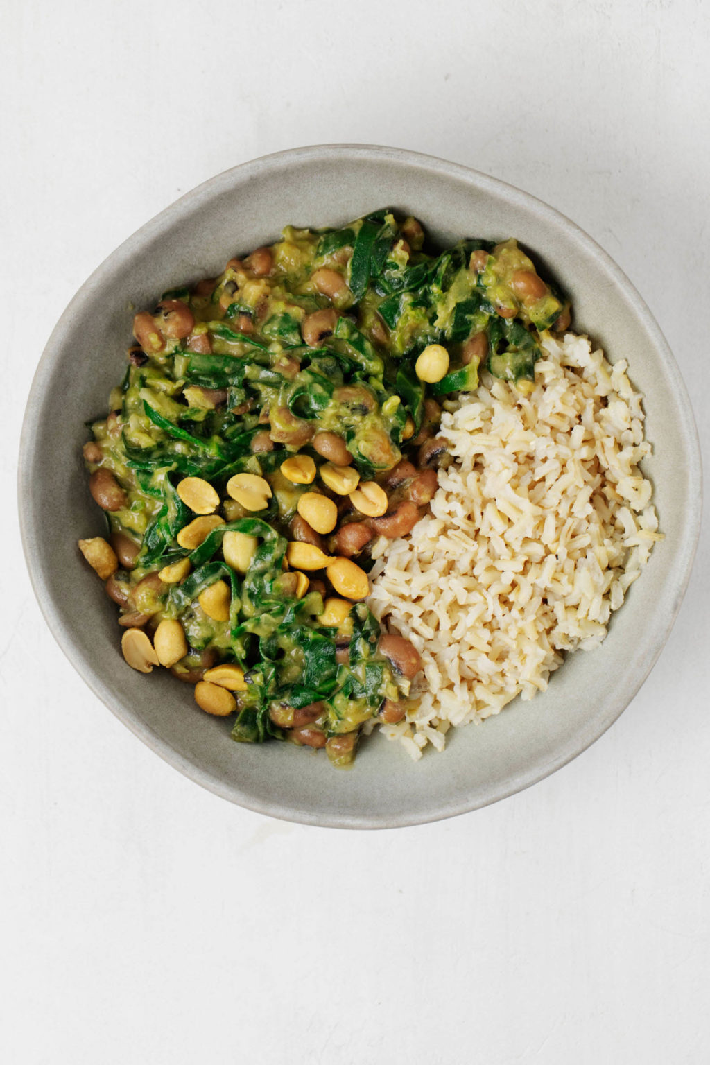 Greens and beans have been served in a ceramic bowl with brown rice. It rests on a white surface.