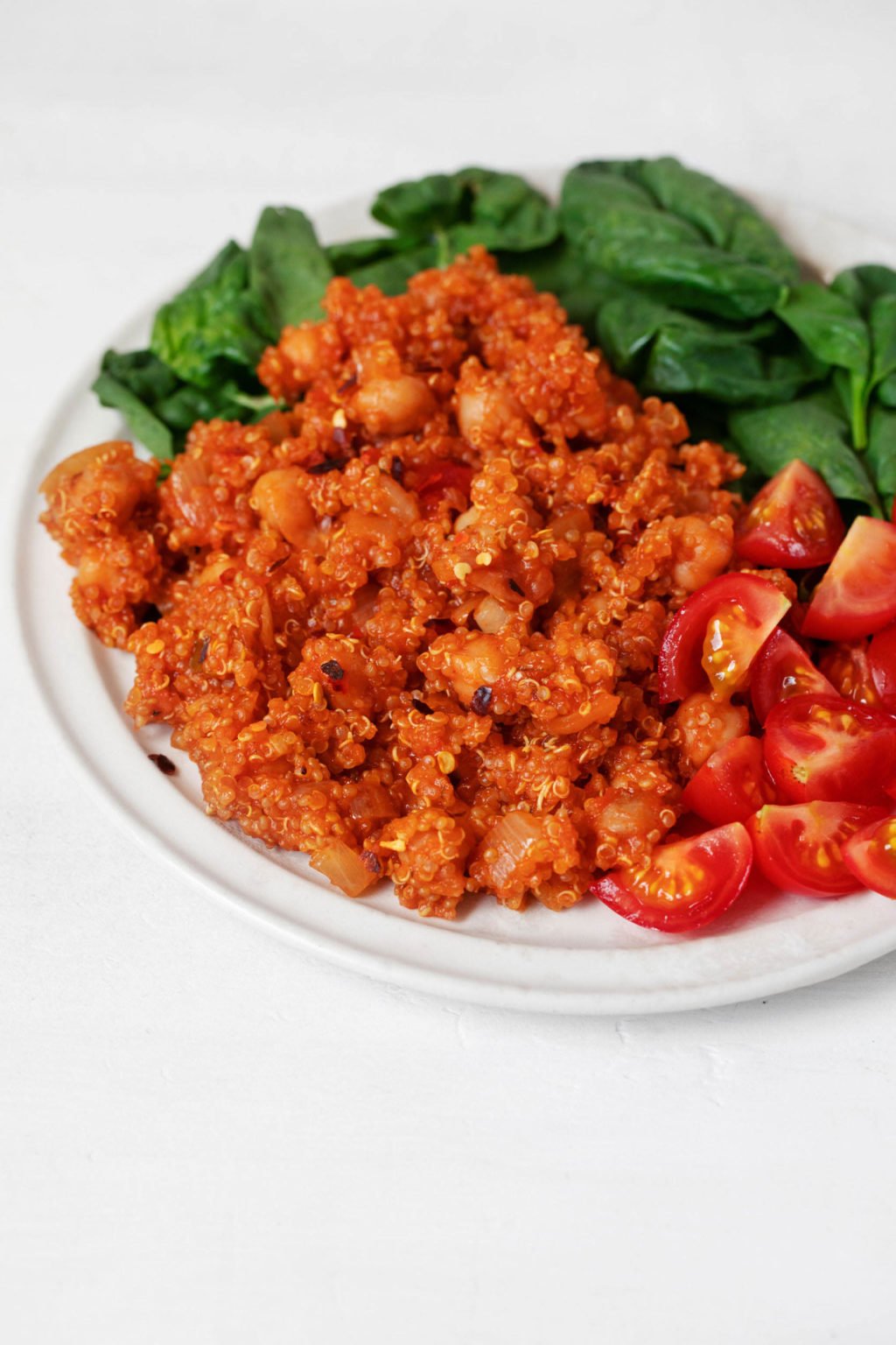 A white, round plate is topped with a tomato-based quinoa and chickpea dish, cherry tomatoes, and baby spinach.