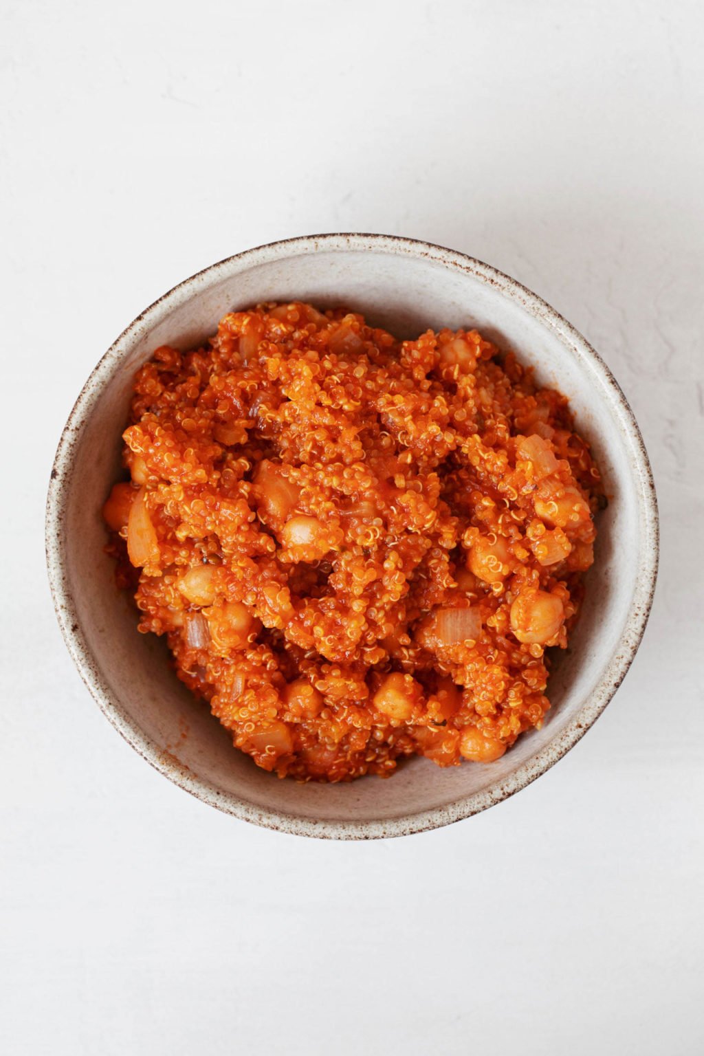 A round, white ceramic bowl is resting on a white surface. It has been filled with an orange-red, seasoned grain pilaf. 