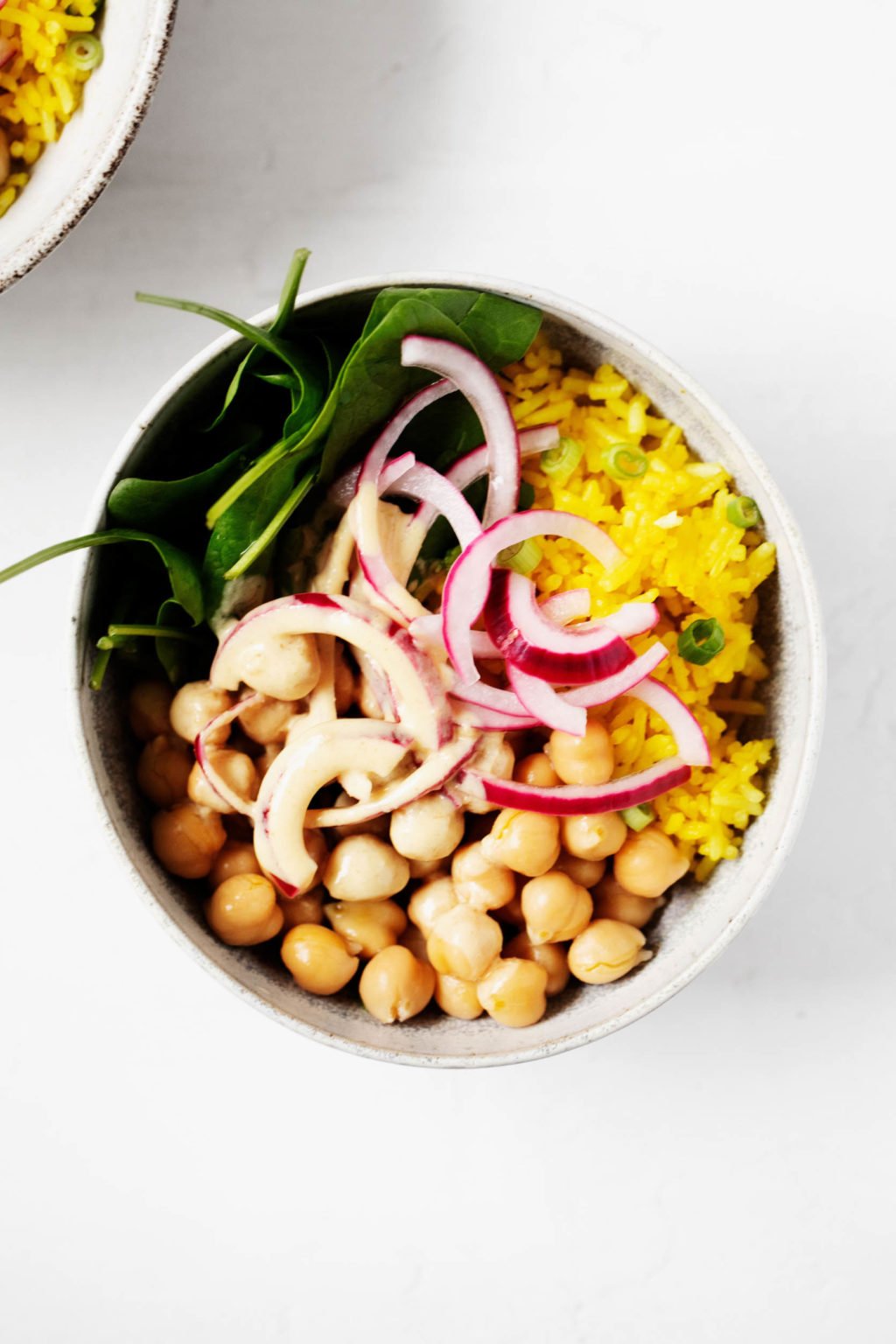 Turmeric rice bowls have been created with chickpeas, turmeric spiced rice, pickled onions, and greens. They're served in two white bowls.