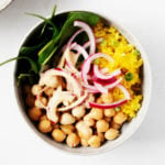 Turmeric rice bowls have been created with chickpeas, turmeric spiced rice, pickled onions, and greens. They're served in two white bowls.