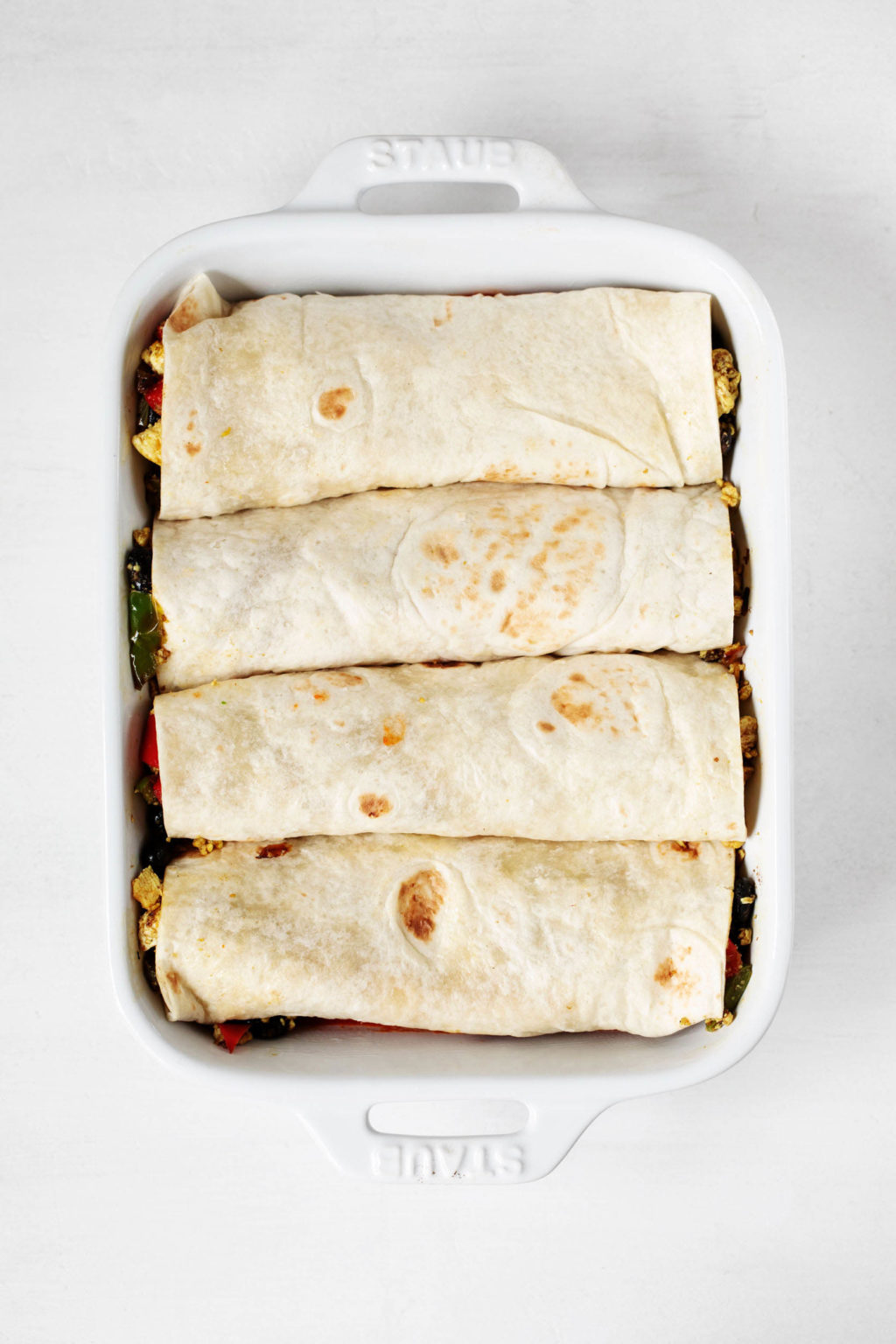 Vegan tortillas, stuffed with a filling, are layered in a white baking dish.