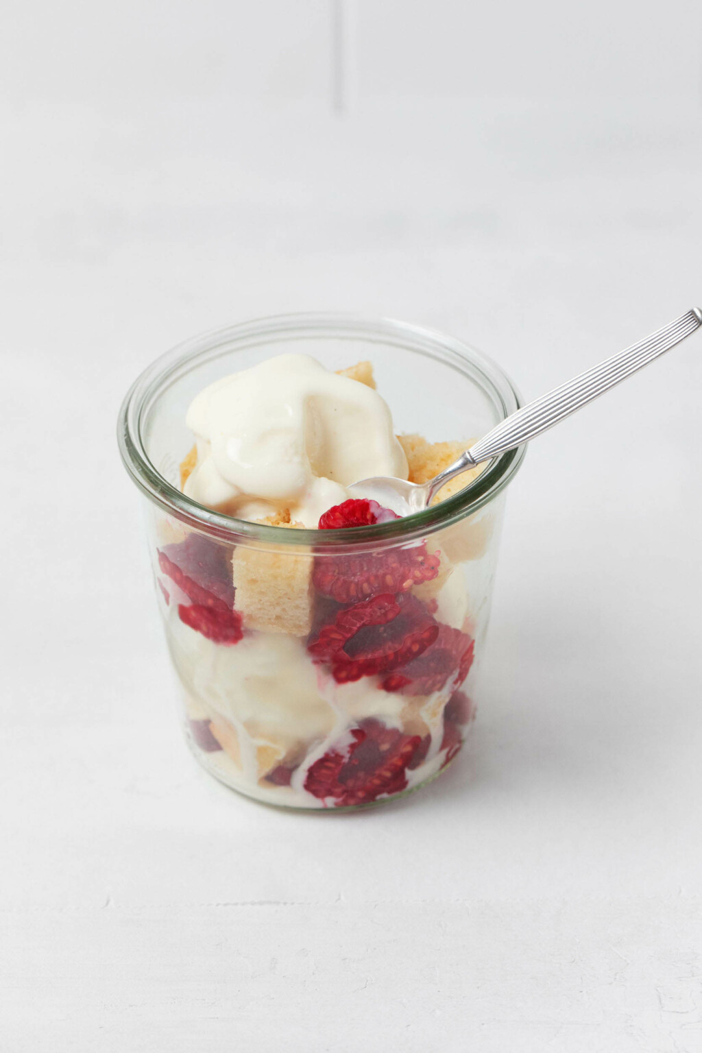 A small, clear glass mason jar holds berries, vegan cake, and vanilla ice cream that's just barely melting. It rests on a white surface.