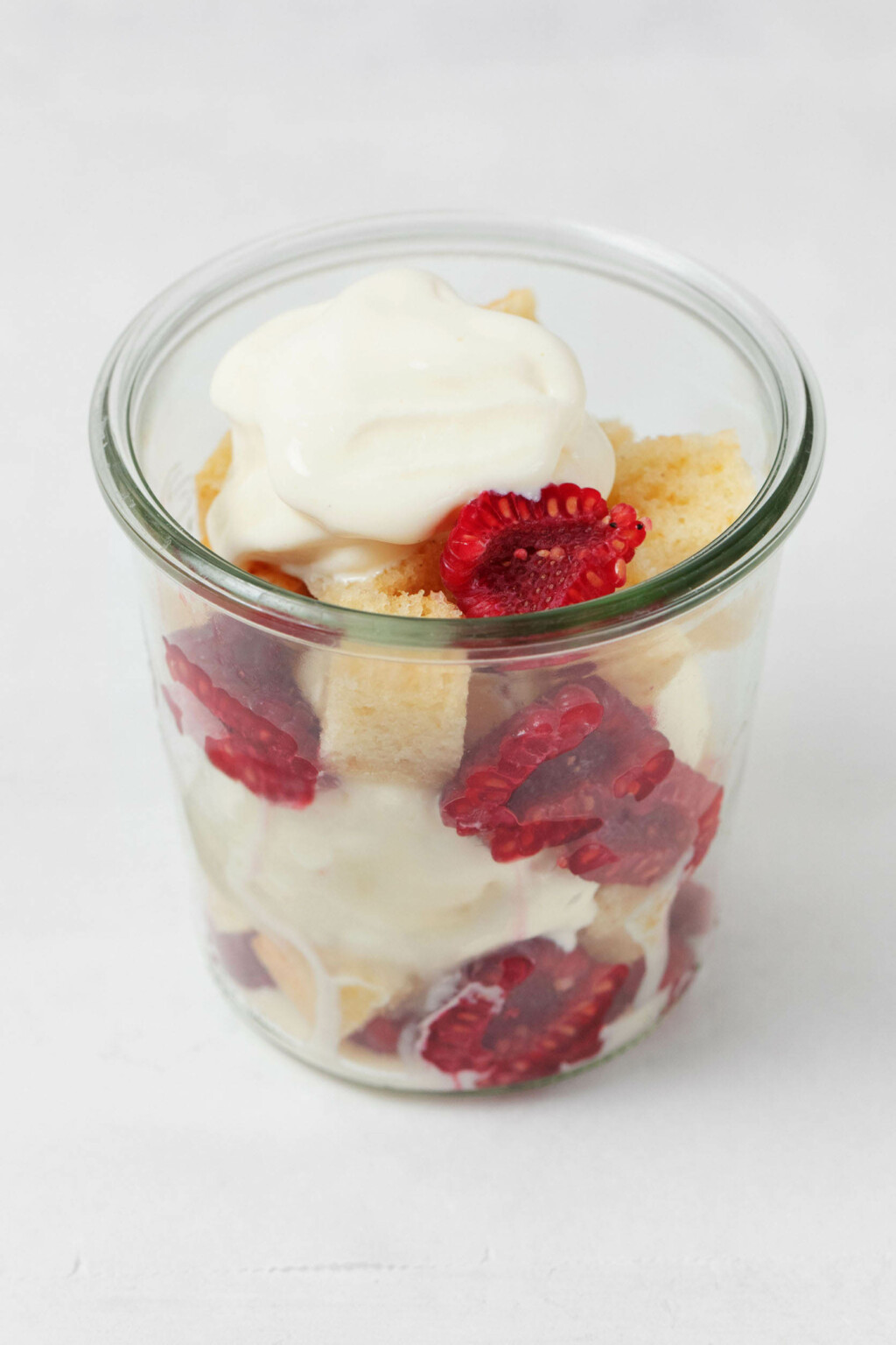 An overhead image of a mason jar that's filled with red raspberries, cubes of lemon cake, and ice cream.
