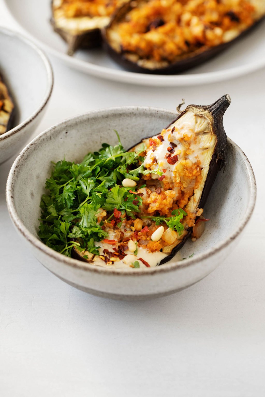 Serving dishes of vegan stuffed eggplant with a bowl in the foreground. Parsley and tahini dressing garnishes.