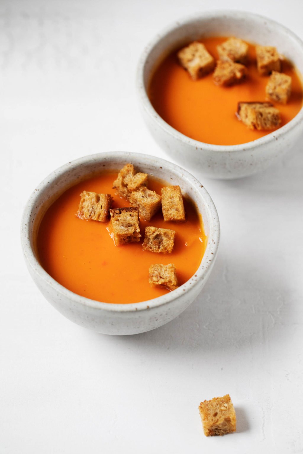 Two bowls of a creamy vegan sweet potato and roasted red pepper soup, topped with homemade croutons.
