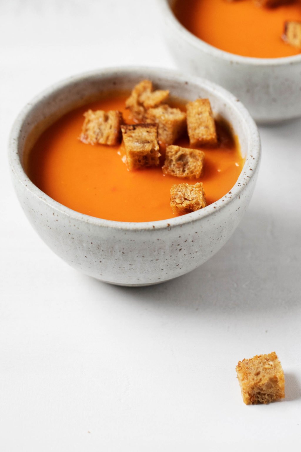 Two bowls of vegan red pepper and potato soup are positioned close to each other, with some rustic bread croutons in the bowls and resting nearby.