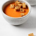 Two bowls of vegan red pepper and potato soup are positioned close to each other, with some rustic bread croutons in the bowls and resting nearby.