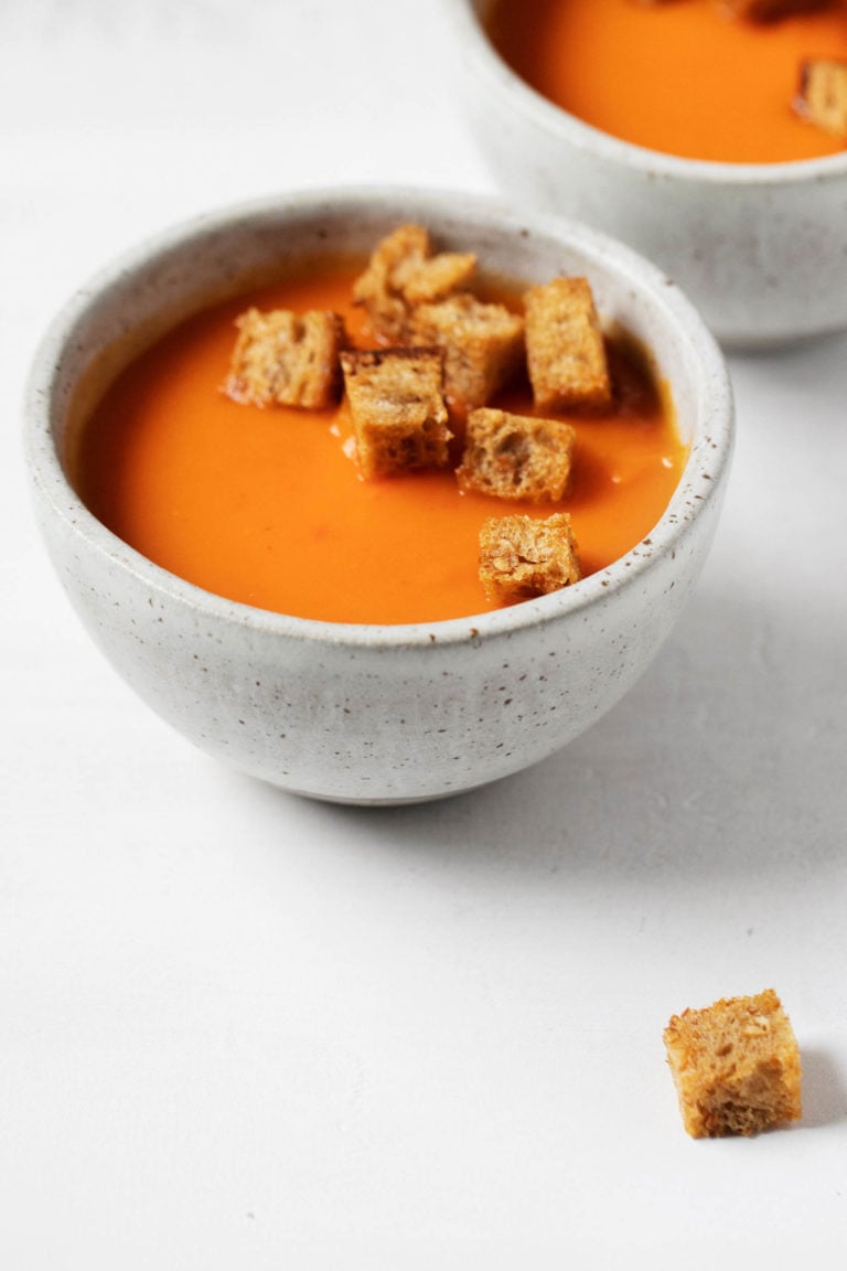 Two bowls of vegan red pepper and potato soup are positioned close to each other, with some rustic bread croutons in the bowls and resting nearby.