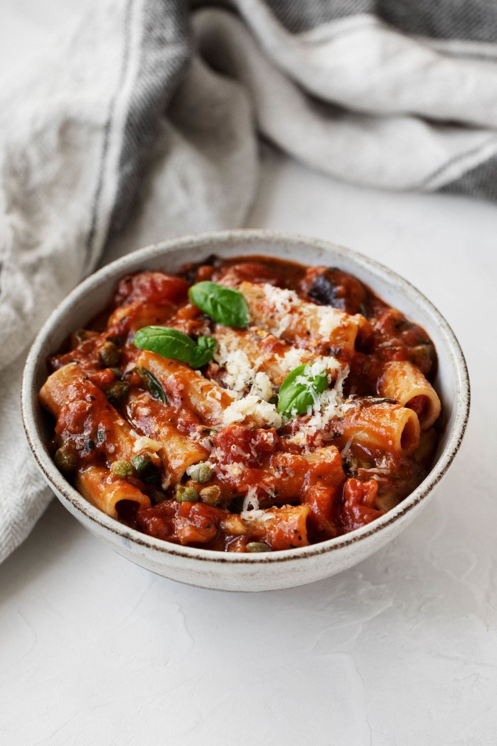 A round bowl contains a dairy-free version of Pasta alla Norma, which has been prepared with eggplant, tomatoes, and capers. A gray linen napkin is positioned nearby.