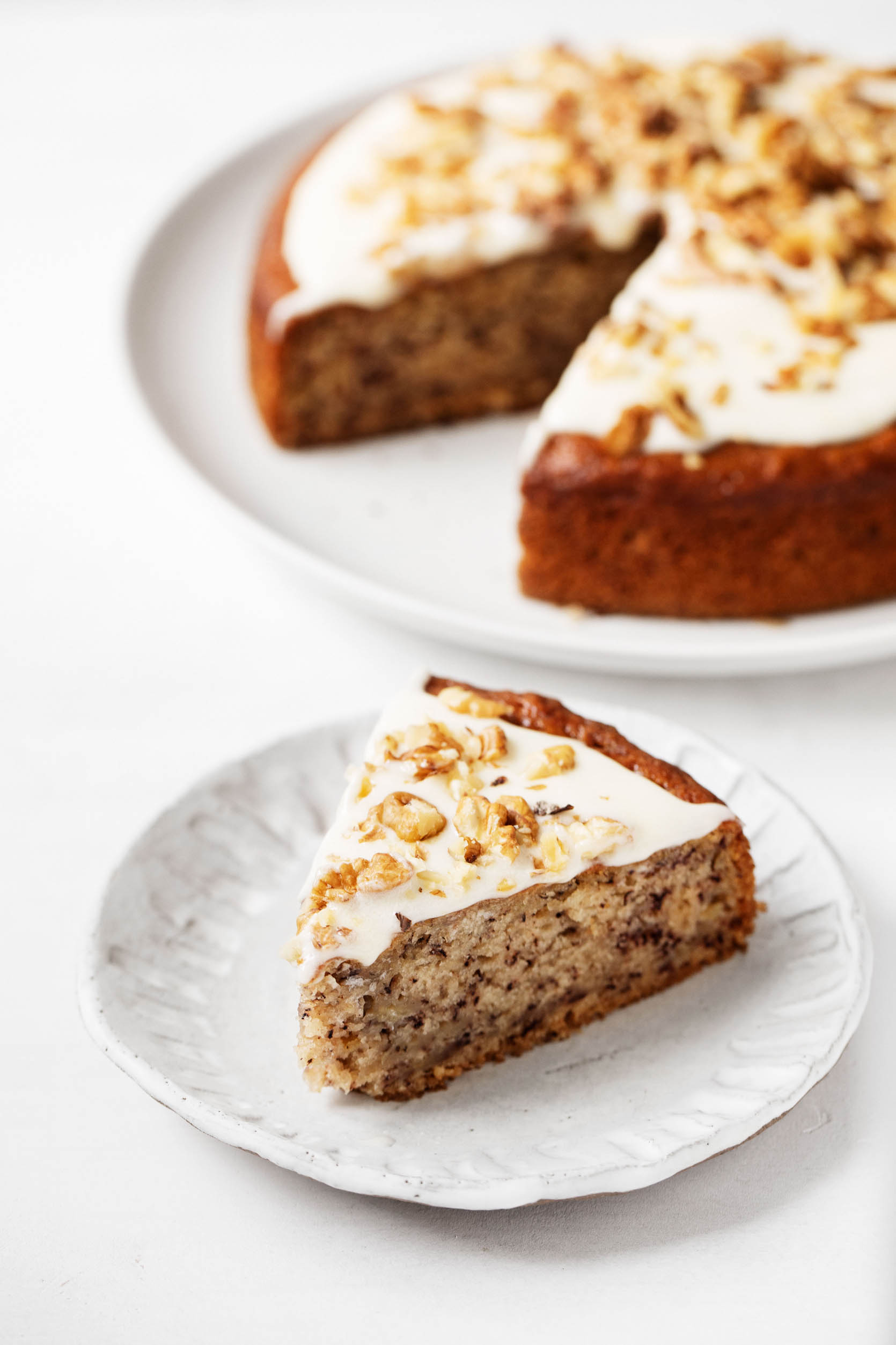 A beautiful, freshly cut slice of vegan banana cake rests on a small dessert plate, decorated with frosting and chopped nuts.