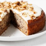 A round vegan banana cake with cashew frosting has been sliced into on a serving plate.