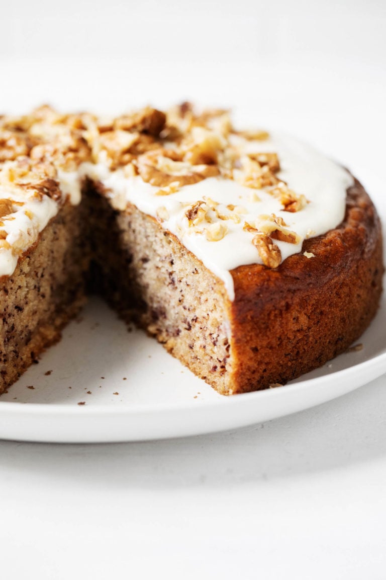 A round vegan banana cake with cashew frosting has been sliced into on a serving plate.