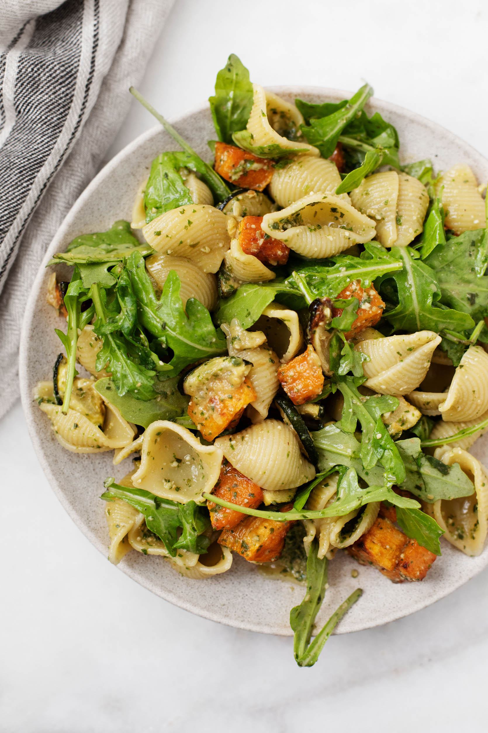 Pasta shells are dressed with freshly made pesto and tossed together with arugula, roasted butternut squash, and zucchini. They rest on a serving plate with a cloth napkin nearby.