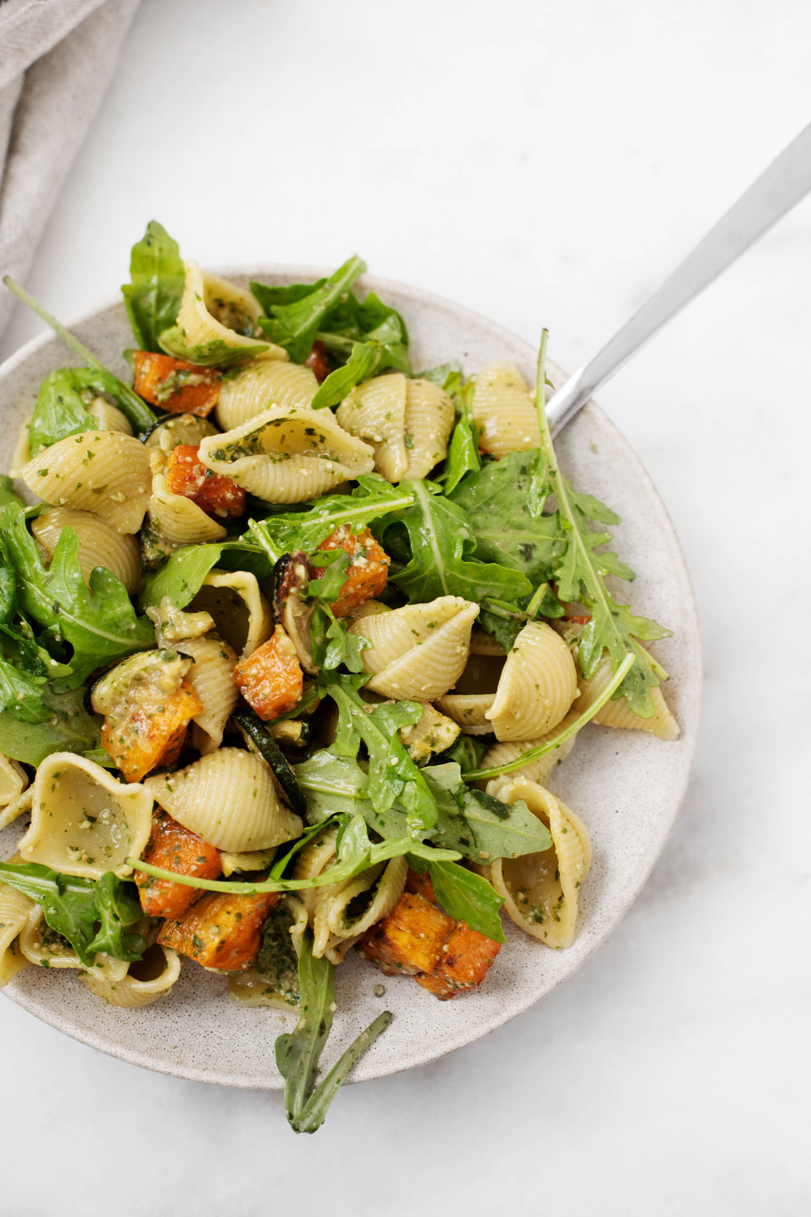 A zoomed in photograph of a plate of pasta shells made with roasted vegetables, arugula, and pesto sauce.