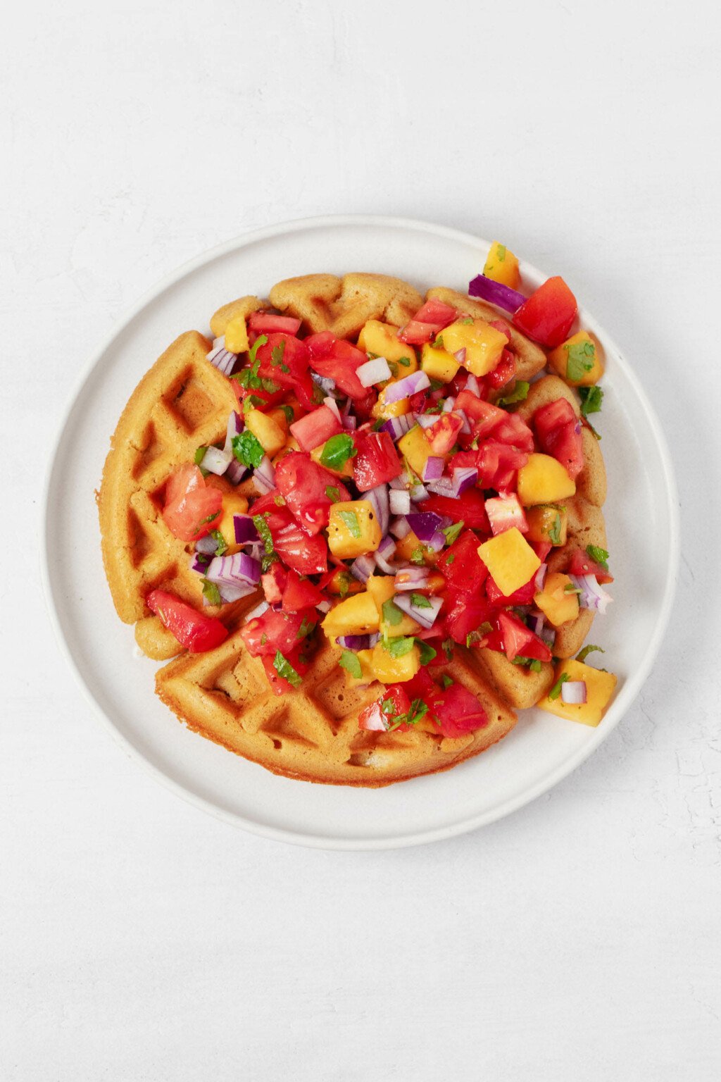 An overhead image of a round vegan cornmeal waffle, which is served on a white plate and topped with tomato peach salsa.