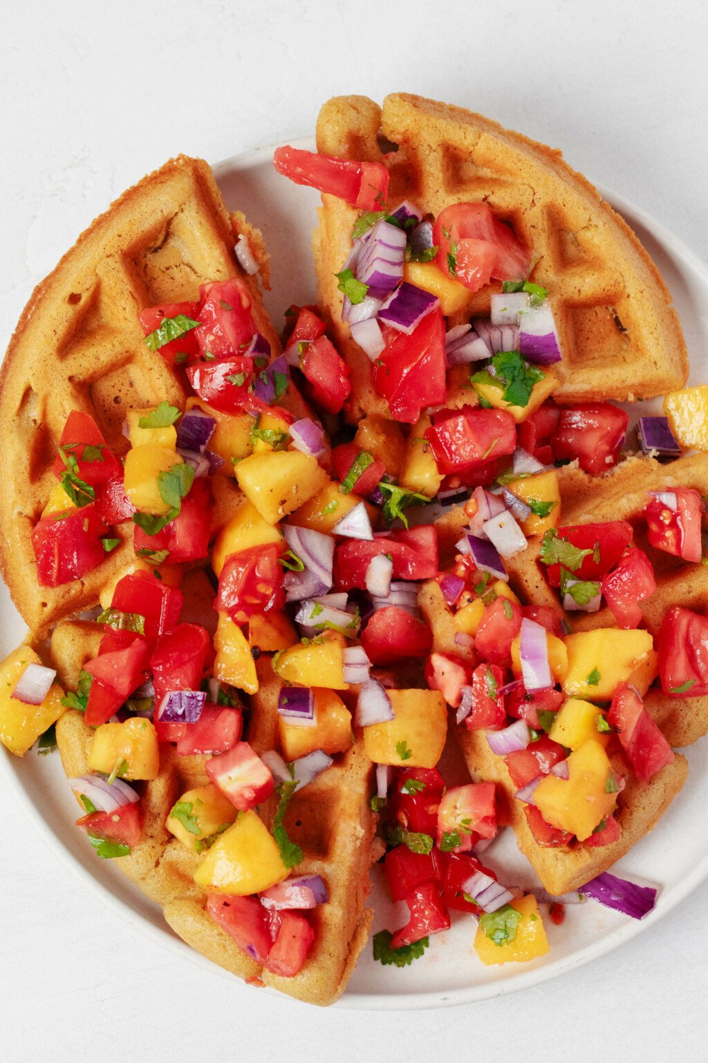 An overhead image of a round vegan cornmeal waffle, which is served on a white plate and topped with tomato peach salsa.