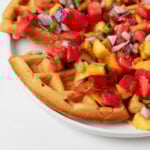 A slice of a vegan crispy cornmeal waffle has been pulled back from the waffle itself. The mixture rests on a white plate.