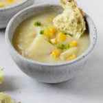 A serving bowl of vegan cauliflower corn chowder is served with a torn piece of bread dunked into the warm soup.