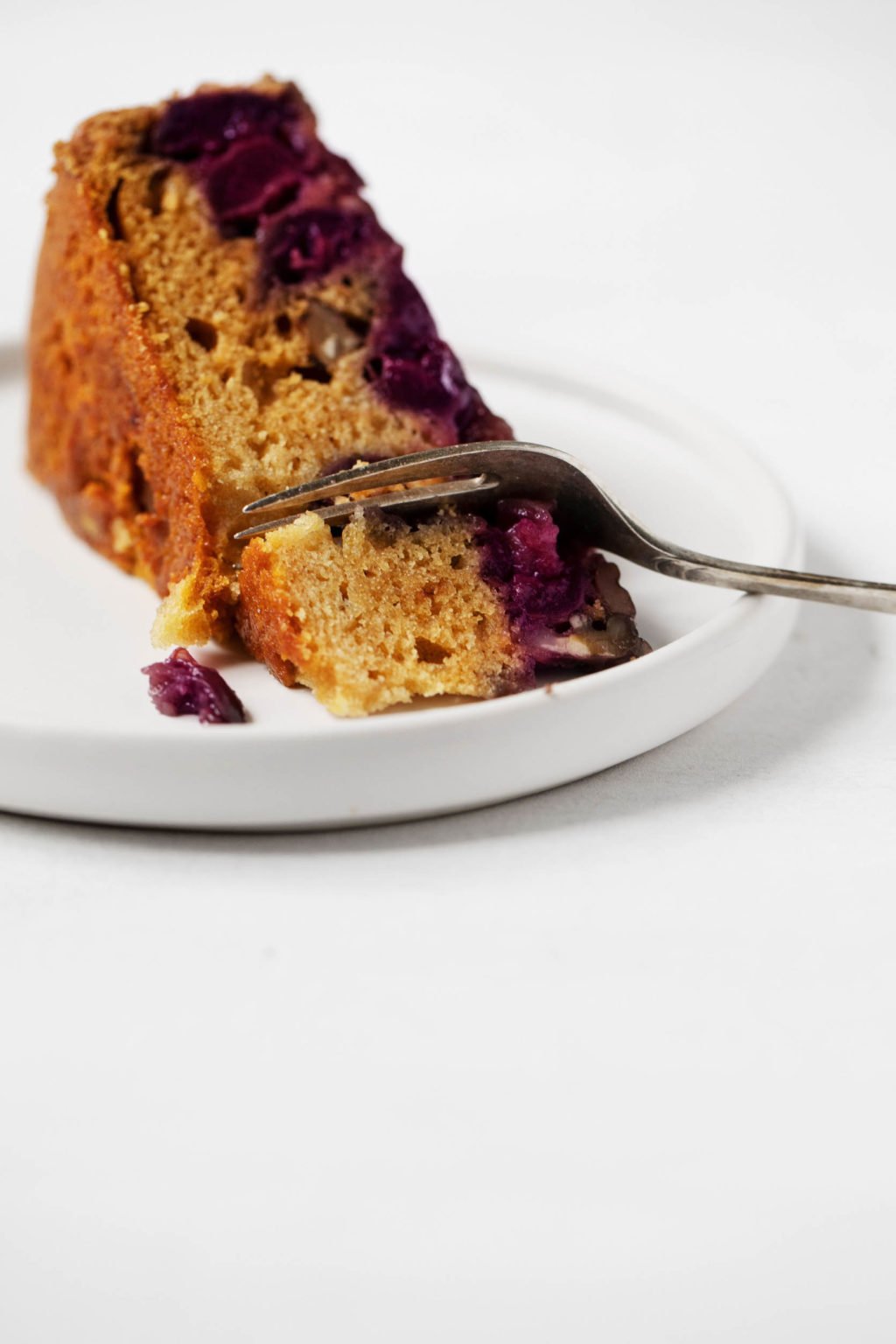 A slice of fruit-topped dessert is positioned on a small white dessert plate and being cut into with a fork.