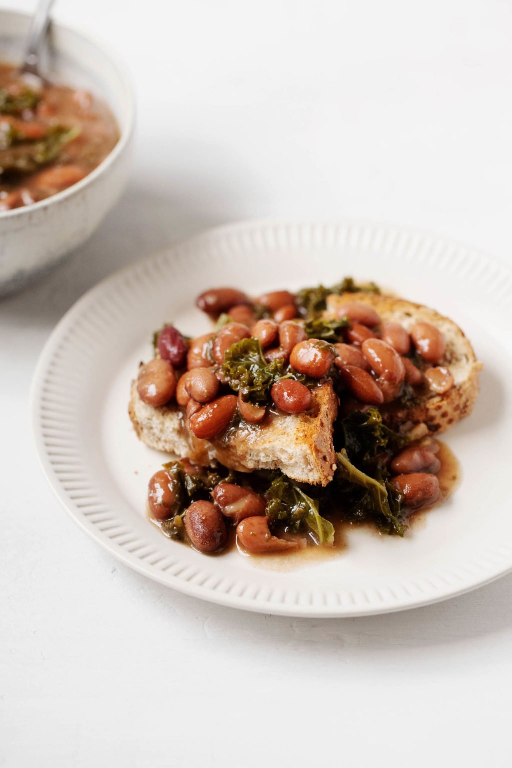 A white serving plate holds toast that has been smothered with braised beans and kale. A bowl with additional ingredients peeks out in the background.