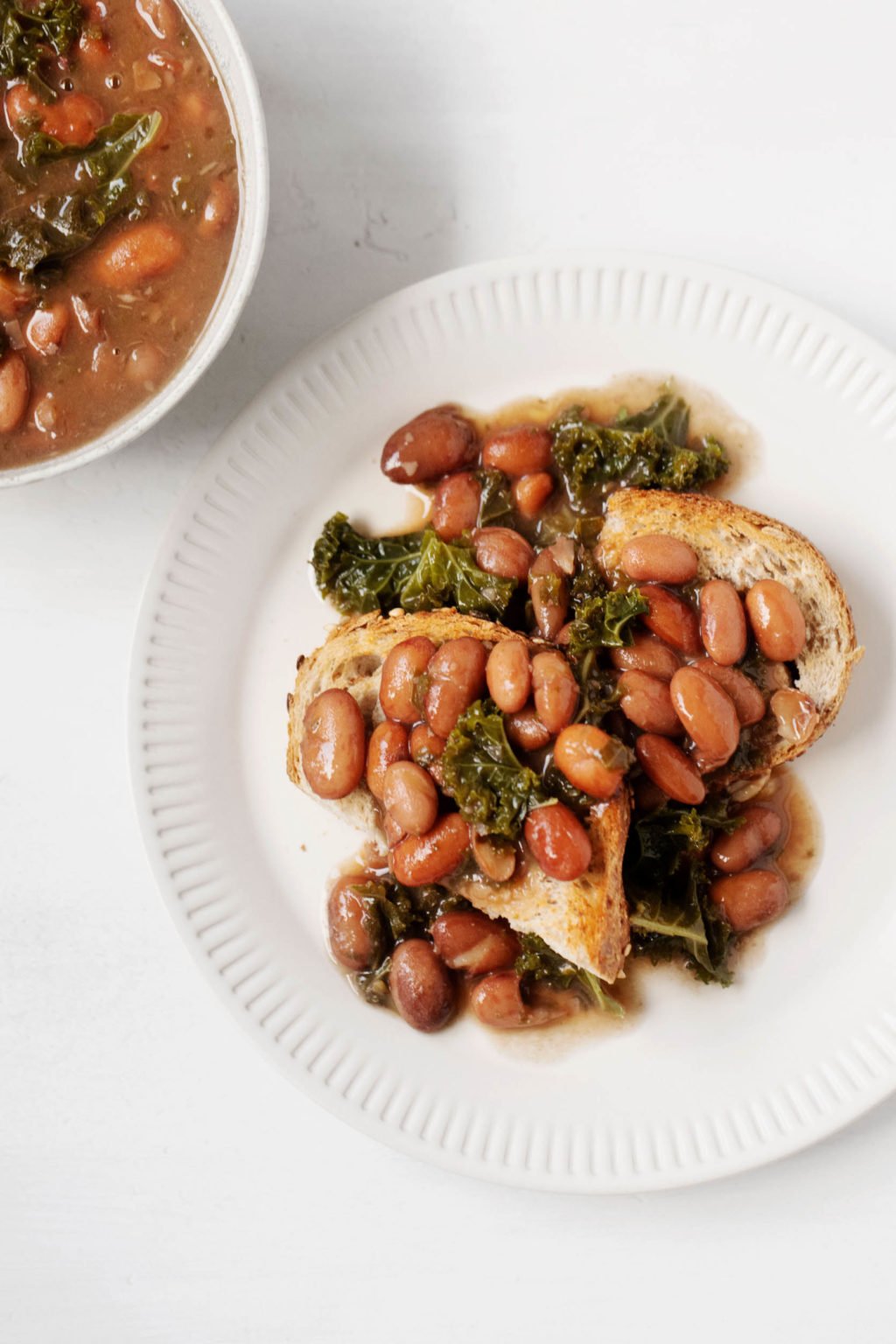A white, ceramic, fluted plate has been covered with a piece of toast and some braised beans & kale.