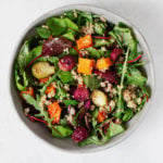 A round, white bowl has been filled with a vibrant roasted butternut squash salad that includes a lot of bright, leafy greens.
