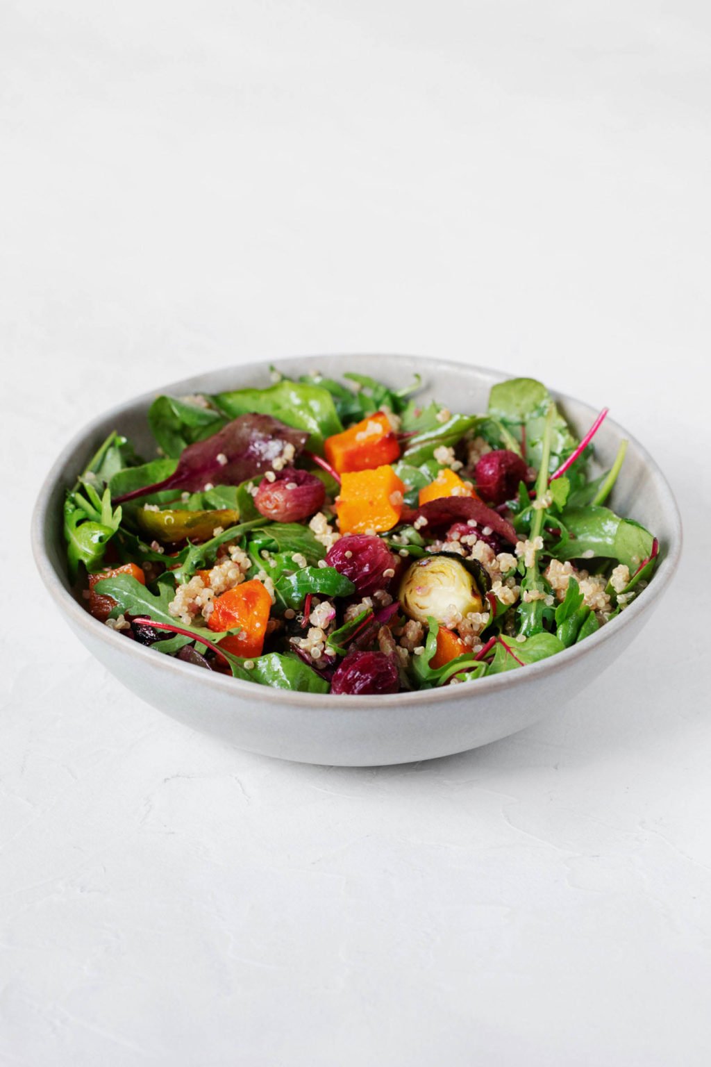 A white, round bowl is resting on a white surface. The bowl is filled with baby greens, cooked winter vegetables, whole grains, and a dressing.