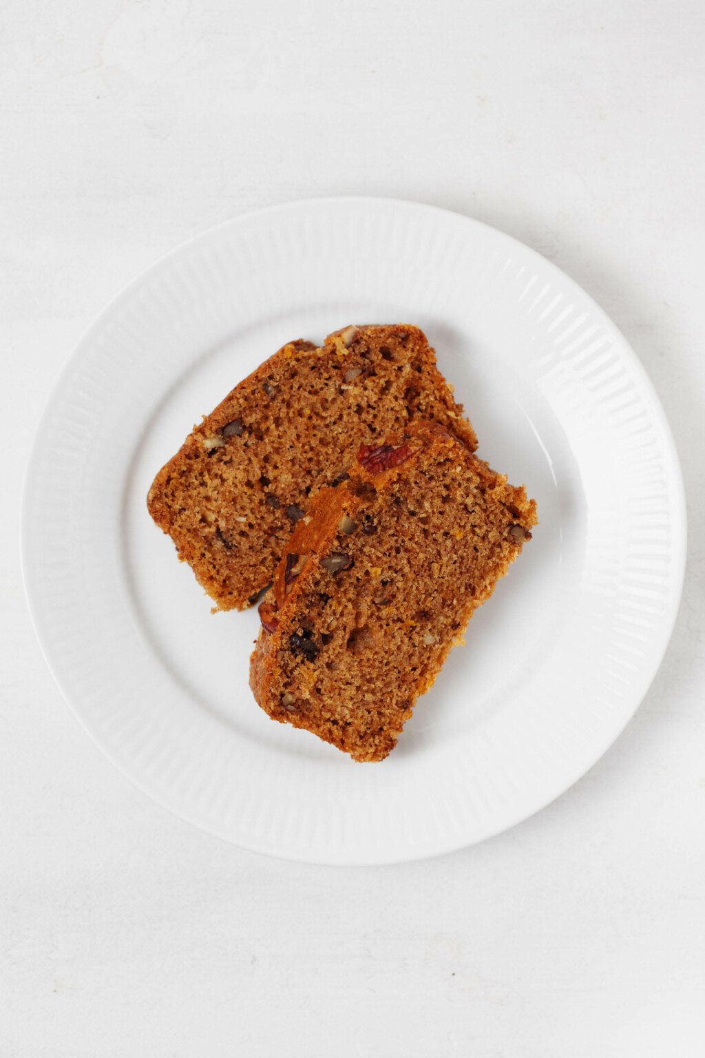 Two slices of vegan spiced pecan citrus loaf are resting on a round, white plate.