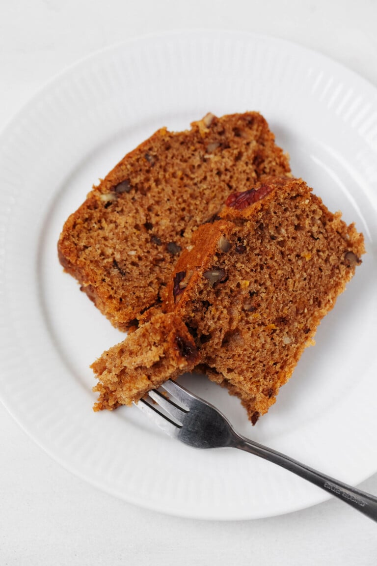 A moist, fluffy vegan pecan citrus loaf has been sliced and plated. A fork is being used to cut into one slice.