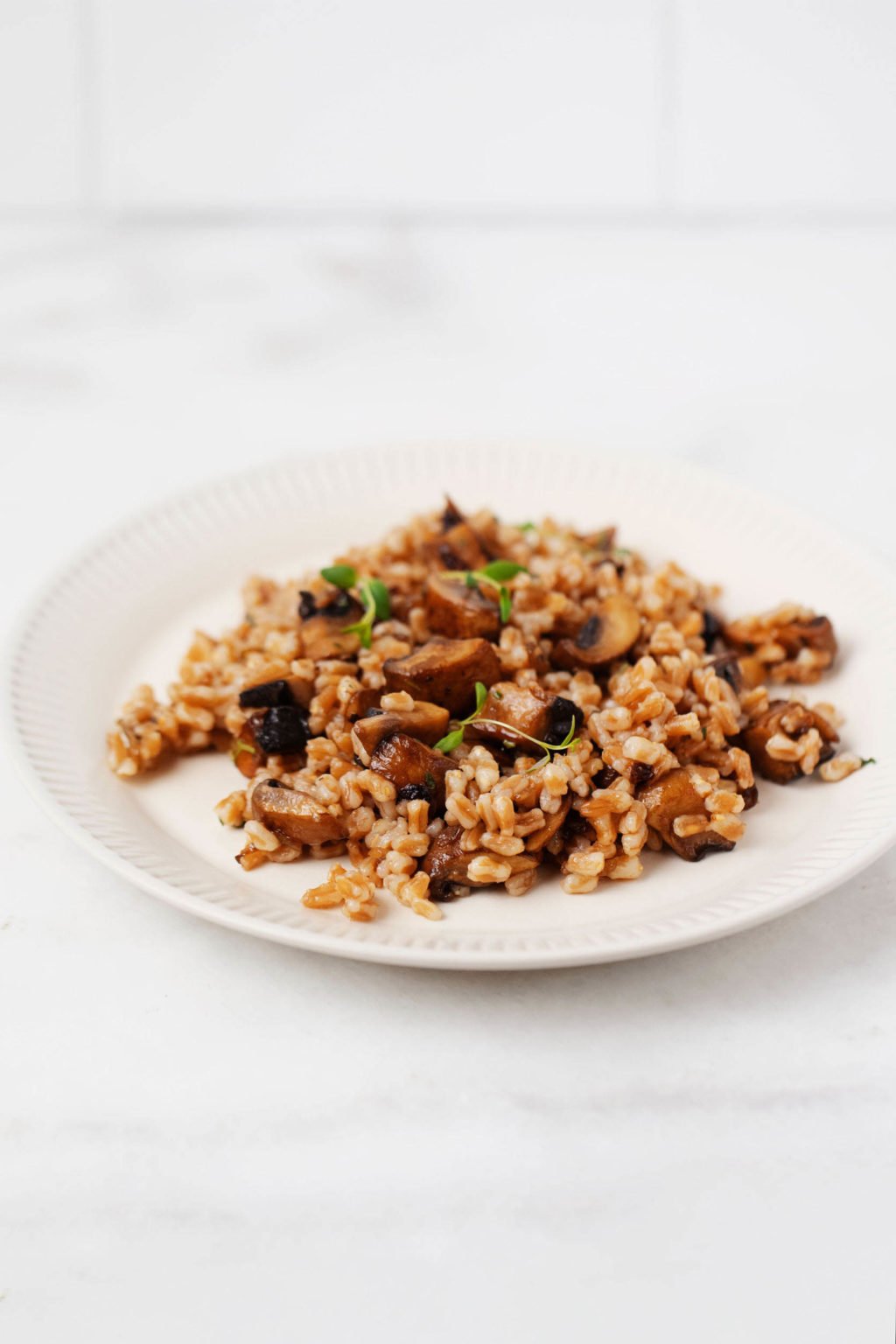 A white, rimmed plate holds a side dish of mushroom farro, topped with herbs. It rests on a white marble surface.