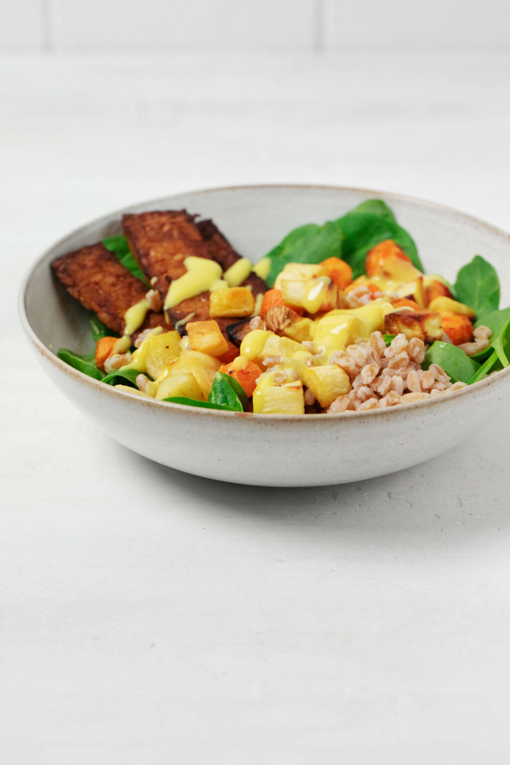 A round, white bowl is resting on a white surface. It's filled with grains, roasted root vegetables, and tempeh.