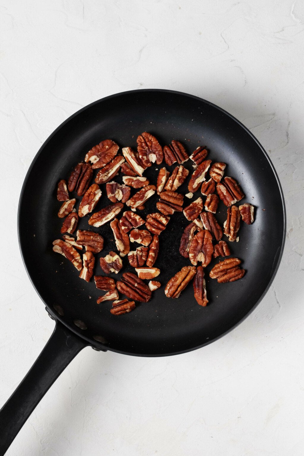 An overhead image of a small, black skillet, filled with toasted pecans.