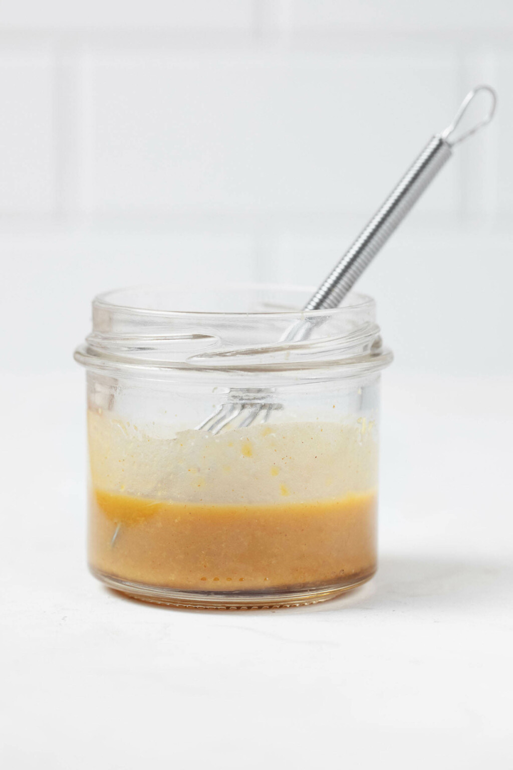 An image of a small mason jar, which is filled with a light-brown colored dressing and a tiny whisk.