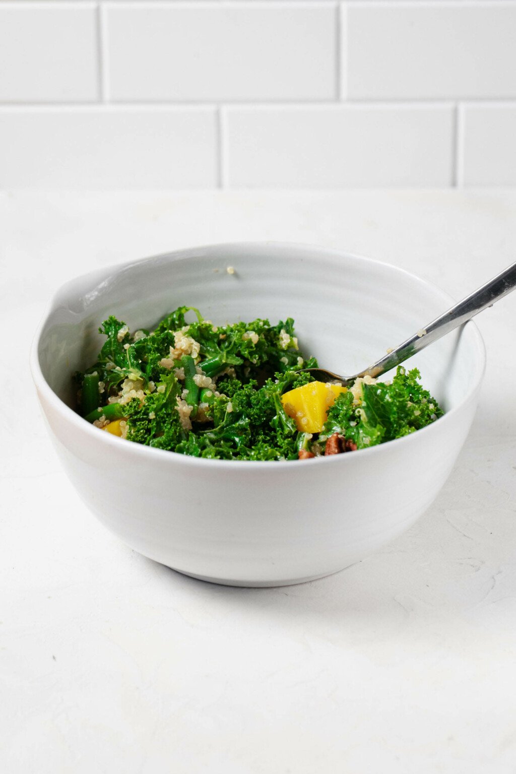 A large, white ceramic mixing bowl is being used to mix a bright green salad of kale, quinoa, and golden beets.