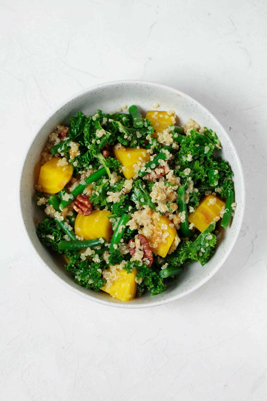 An overhead image of a white bowl, which has been filled with a colorful kale & quinoa salad. The salad also contains roasted beets and green beans.