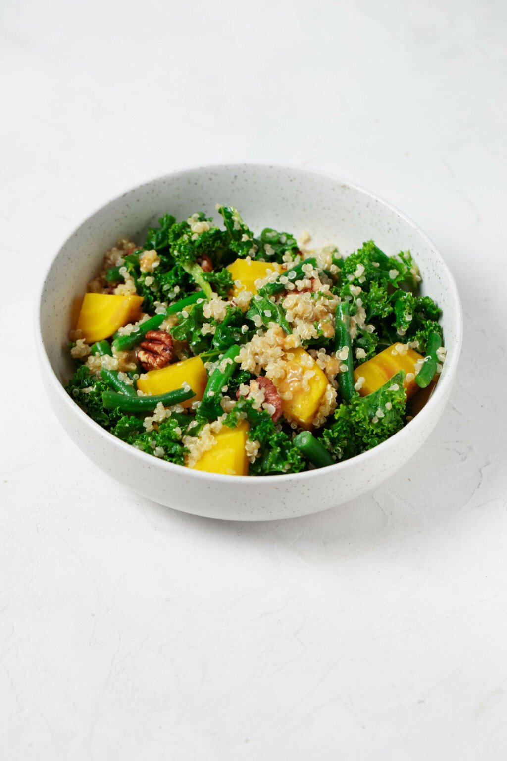 An image of a white bowl, which is filled with a vibrantly-colored kale, golden beet, green bean, and quinoa salad. There are brown pieces of toasted pecan throughout.