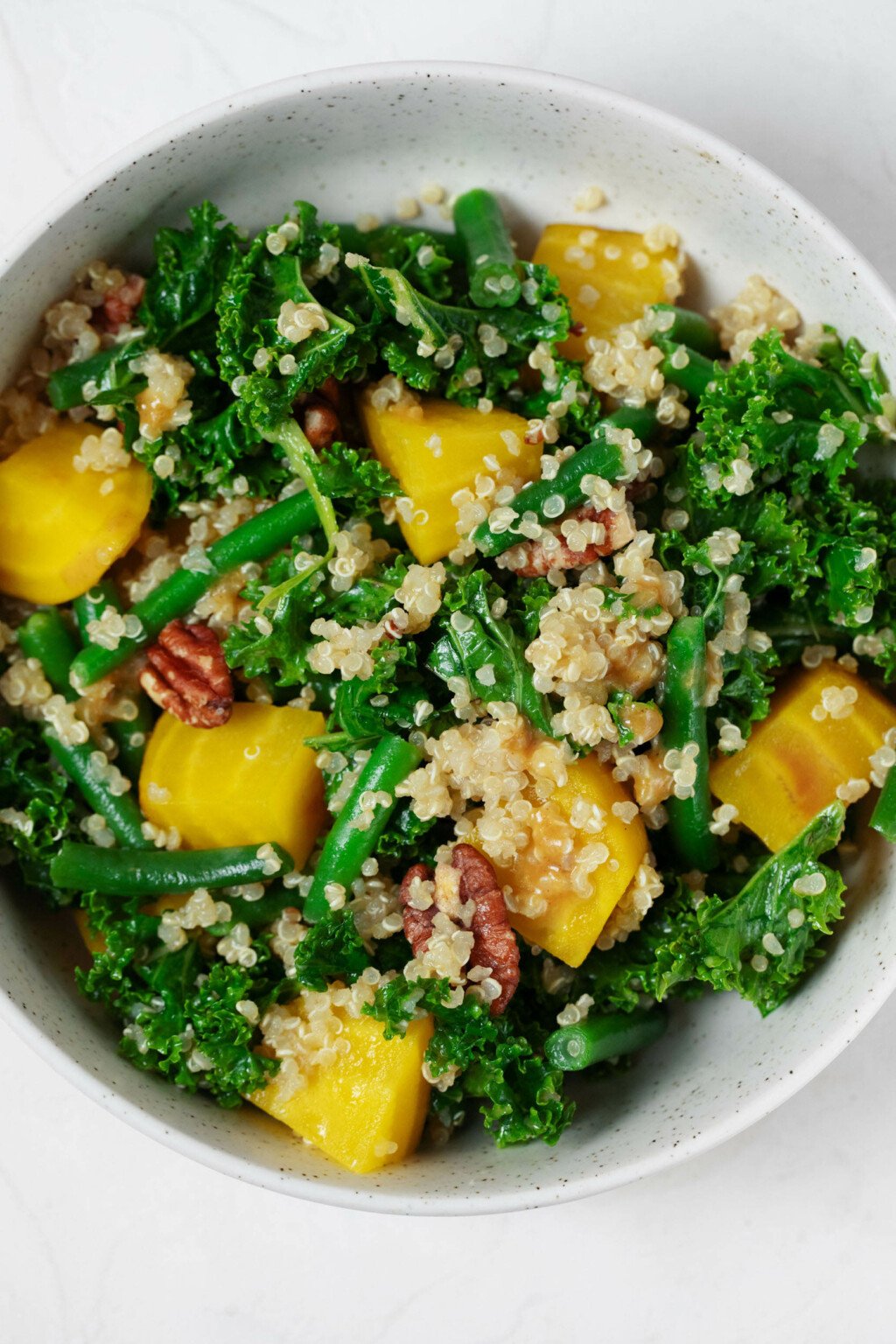 An overhead image of a white bowl, which is filled with a vibrantly-colored kale, golden beet, green bean, and quinoa salad. There are brown pieces of toasted pecan throughout.