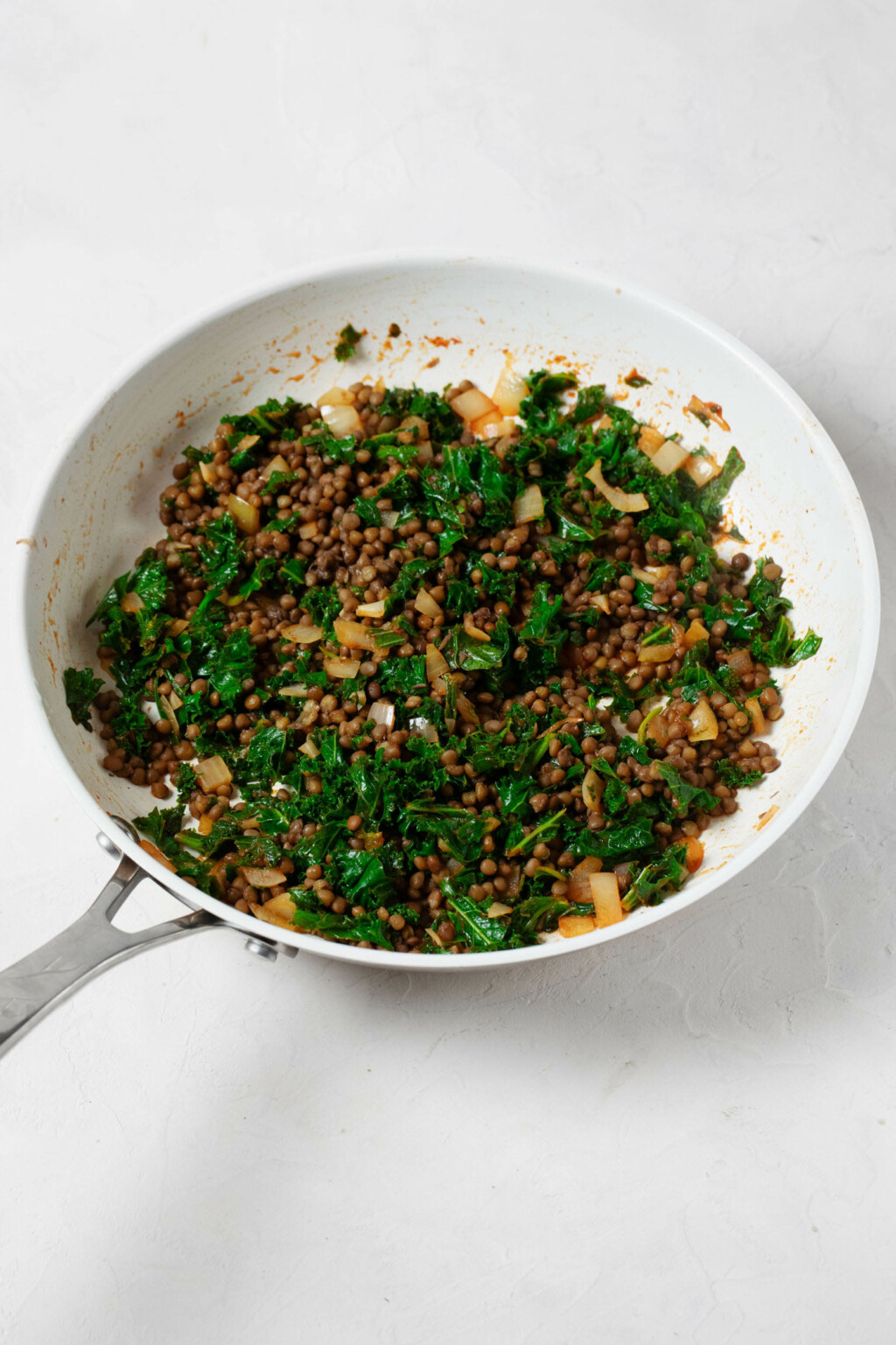 A white, non-stick sauté pan is being used to cook a mixture of brown lentils and green kale.