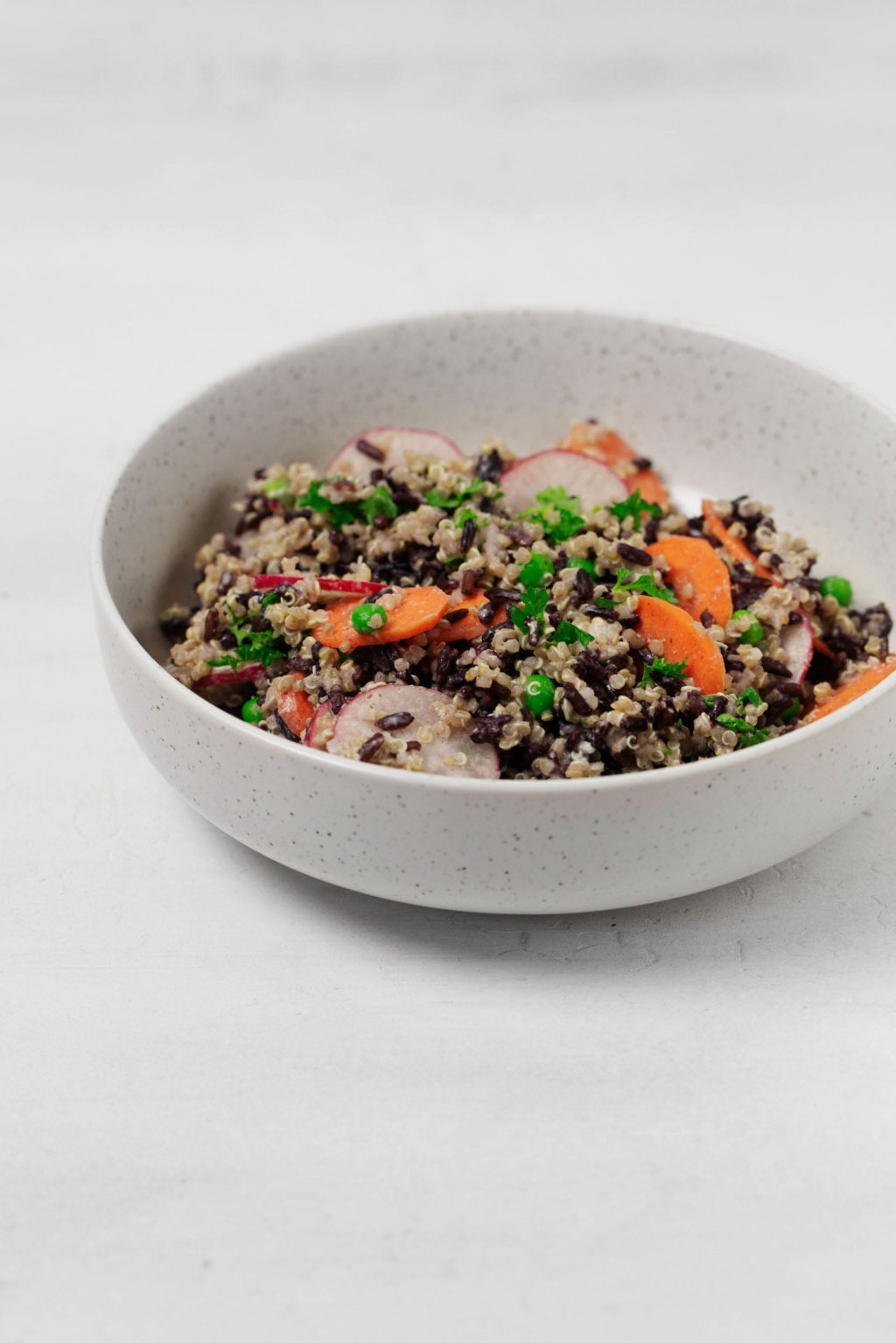 An angled image of a round white bowl, which is filled with a creamy, whole grain quinoa black rice salad.