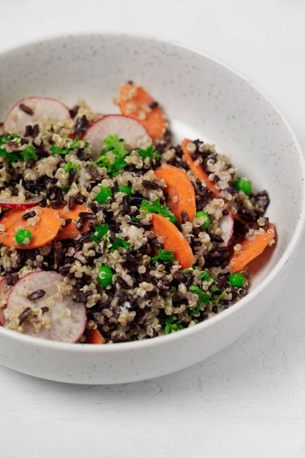 A round, white bowl is resting on a white surface. It's filled with a creamy whole grain and vegetable salad.