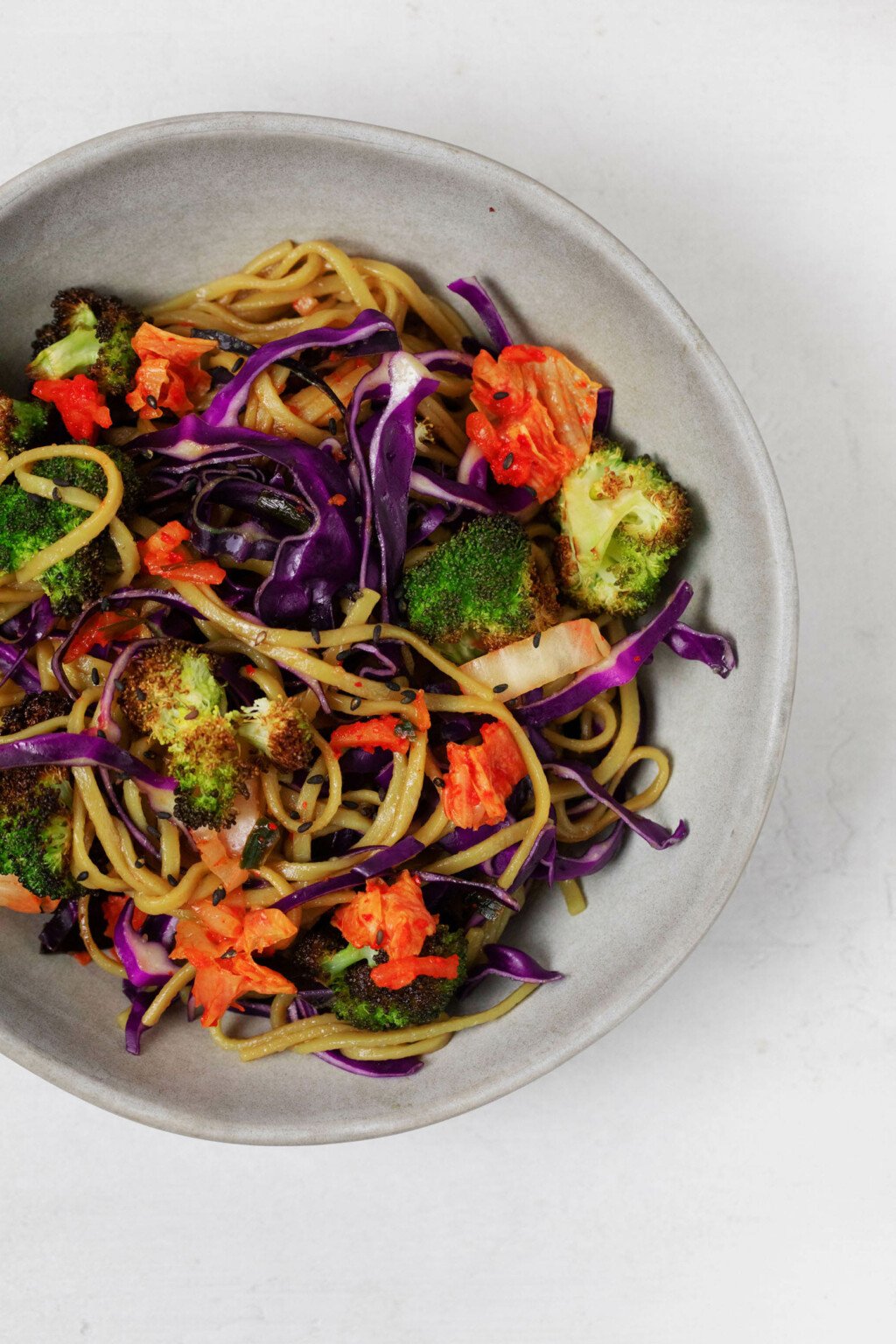 An overhead image of a bright, colorful mix of noodles, roasted broccoli, and crunchy red cabbage, all plated in a gray ceramic bowl.