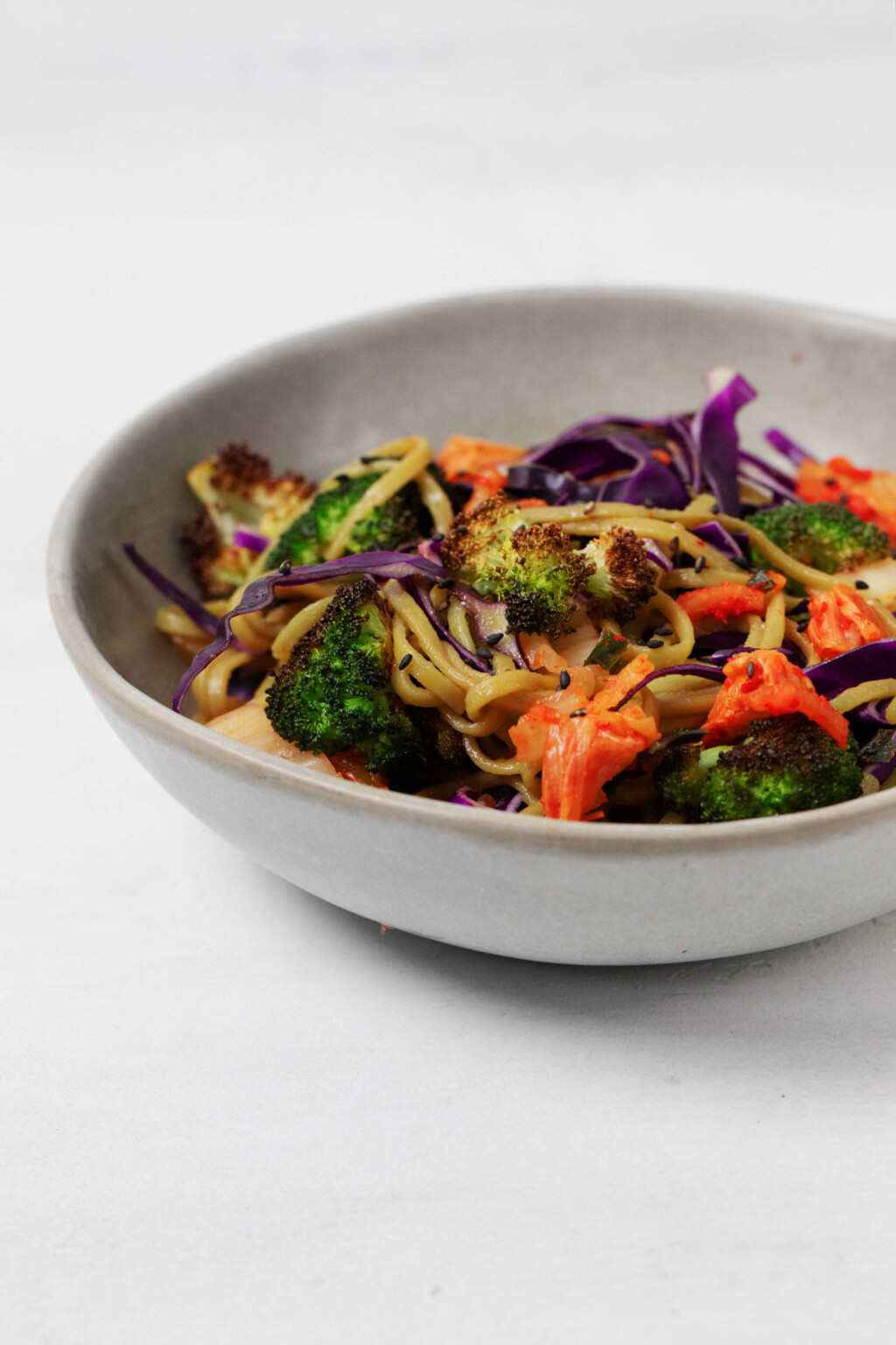 A gray ceramic bowl holds cooked noodles and a bright mixture of raw and cooked vegetables. It rests on a white surface.